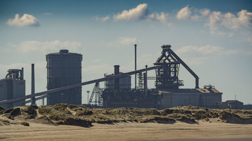 Cranes in factory by sea against sky