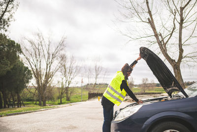 Young caucasian woman looking at car engine, car broken down in the middle of the road. 