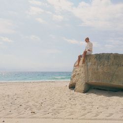 Low angle view of man sitting on rock at beach against cloudy sky