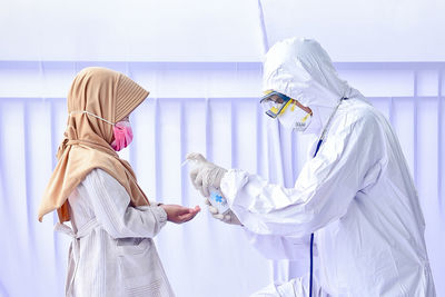 Doctor wearing protective suit giving hand sanitizer to girl against white background