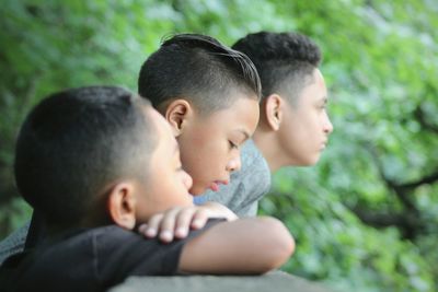 Siblings looking away while leaning on retaining wall against tree at park