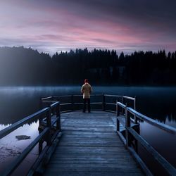 Rear view of man standing on pier over lake against sky during sunset