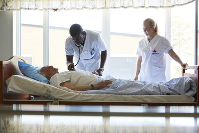 Female nurse looking at male colleague examining senior man lying on bed in hospital ward