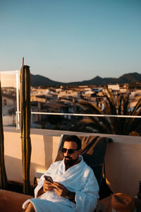 Front view of young bearded male traveler in white robe and sunglasses relaxing on lounger on rooftop and messaging on smartphone against sunset sky during vacation in mallorca