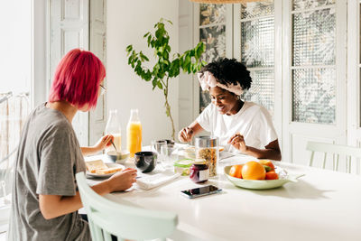 Young multiethnic women sitting at table and eating healthy food during breakfast in sunny morning in modern apartment