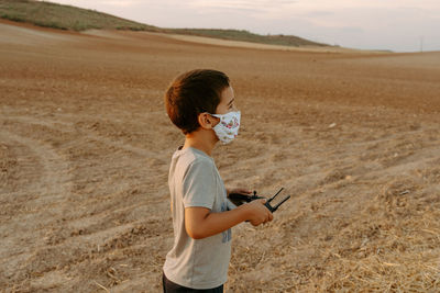 Side view of focused little asian boy in protective mask using remote control while playing with flying drone in field
