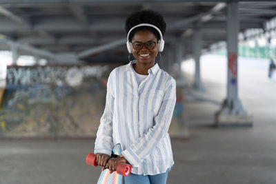 Trendy african skater girl in space for skateboarding wearing wireless headphones and happy smiling