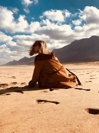 Woman sitting on sand at beach against cloudy sky