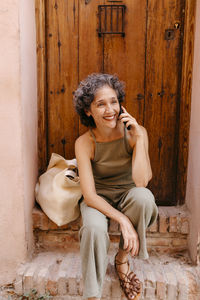 Mature woman talking on smart phone while sitting on doorway