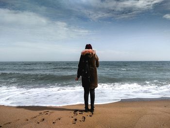 Rear view of young woman in warm clothing standing at beach against sky during sunny day