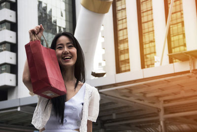 Cheerful woman holding shopping bag while standing against building