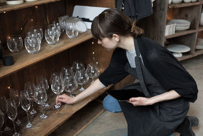 High angle view of young female owner with tablet arranging wineglass while crouching by rack at store