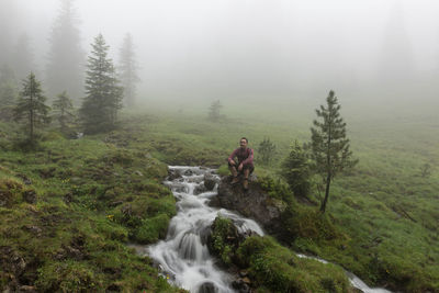 Man sitting by stream on rock at field during foggy weather