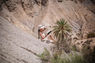 Woman wearing hat reading book while sitting against mountain