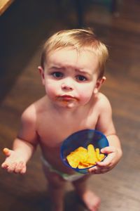 High angle portrait of cute shirtless baby boy with snack standing on hardwood floor at home