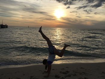 Young woman doing handstand on shore against sky during sunset