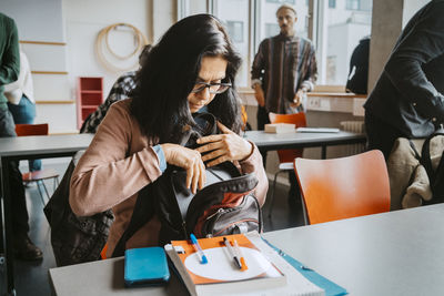 Mature female university student removing books from backpack while sitting at desk in classroom