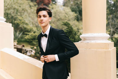 A beautiful young man, the groom in an elegant wedding suit, stands posing in the city's old park