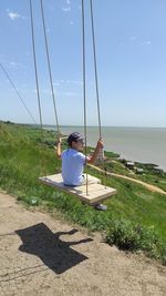 Rear view of child sitting on swing at sea shore against sky