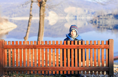 Portrait of boy in warm clothing standing behind wooden railing against calm lake