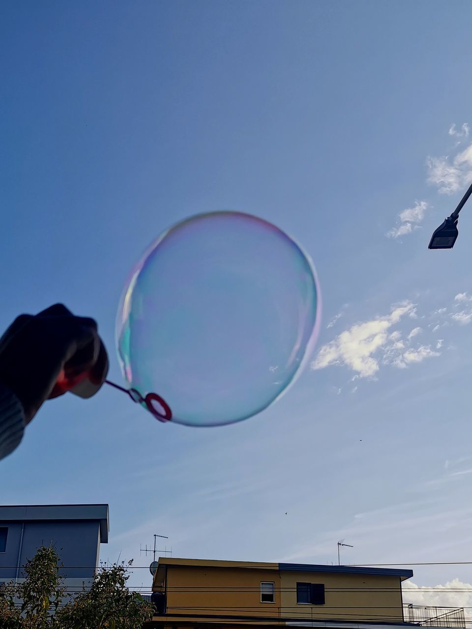 bubble, mid-air, soap sud, sky, bubble wand, fragility, flying, nature, toy, one person, blowing, blue, day, liquid bubble, transparent, motion, architecture, outdoors, low angle view, built structure, building exterior, holding, light, lighting, cloud, men, lightweight, shape, sphere, plant, clear sky, multi colored, environment