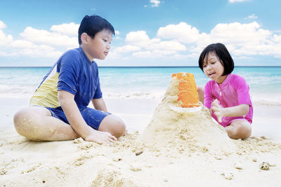 Siblings making sandcastle at beach against cloudy sky