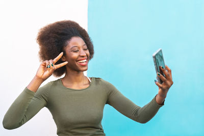 Cheerful young woman taking selfie with mobile phone against wall