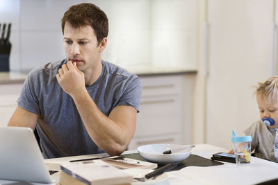 Father working on laptop while baby boy sitting at dining table