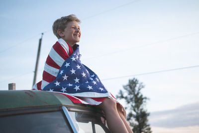 Happy boy with american flag sitting on vehicle against sky during sunset