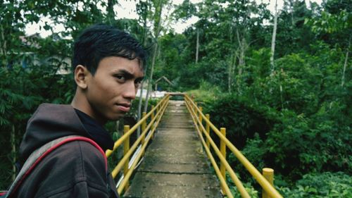 Portrait of teenage boy standing on footbridge amidst trees in forest