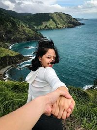 Cropped image of man holding woman while standing on cliff at sea against sky