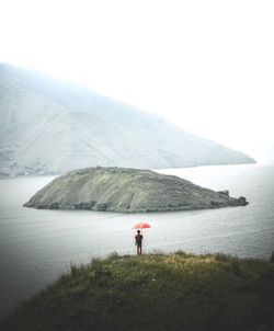 Rear view of man holding umbrella standing on mountain by lake against sky