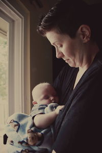 Side view of lesbian woman carrying baby while standing by window at home