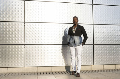 Serious african american male model in fancy outfit standing near silver wall on street and looking at camera