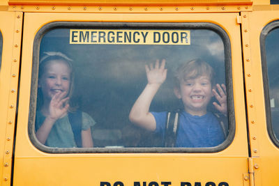 Funny happy smiling boy and girl kids students looking out of school yellow bus window. 
