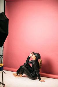 Side view of full body barefoot hispanic lady with ponytail sitting on floor in studio. she is posing in black leather clothes and cap near pink wall and looking up
