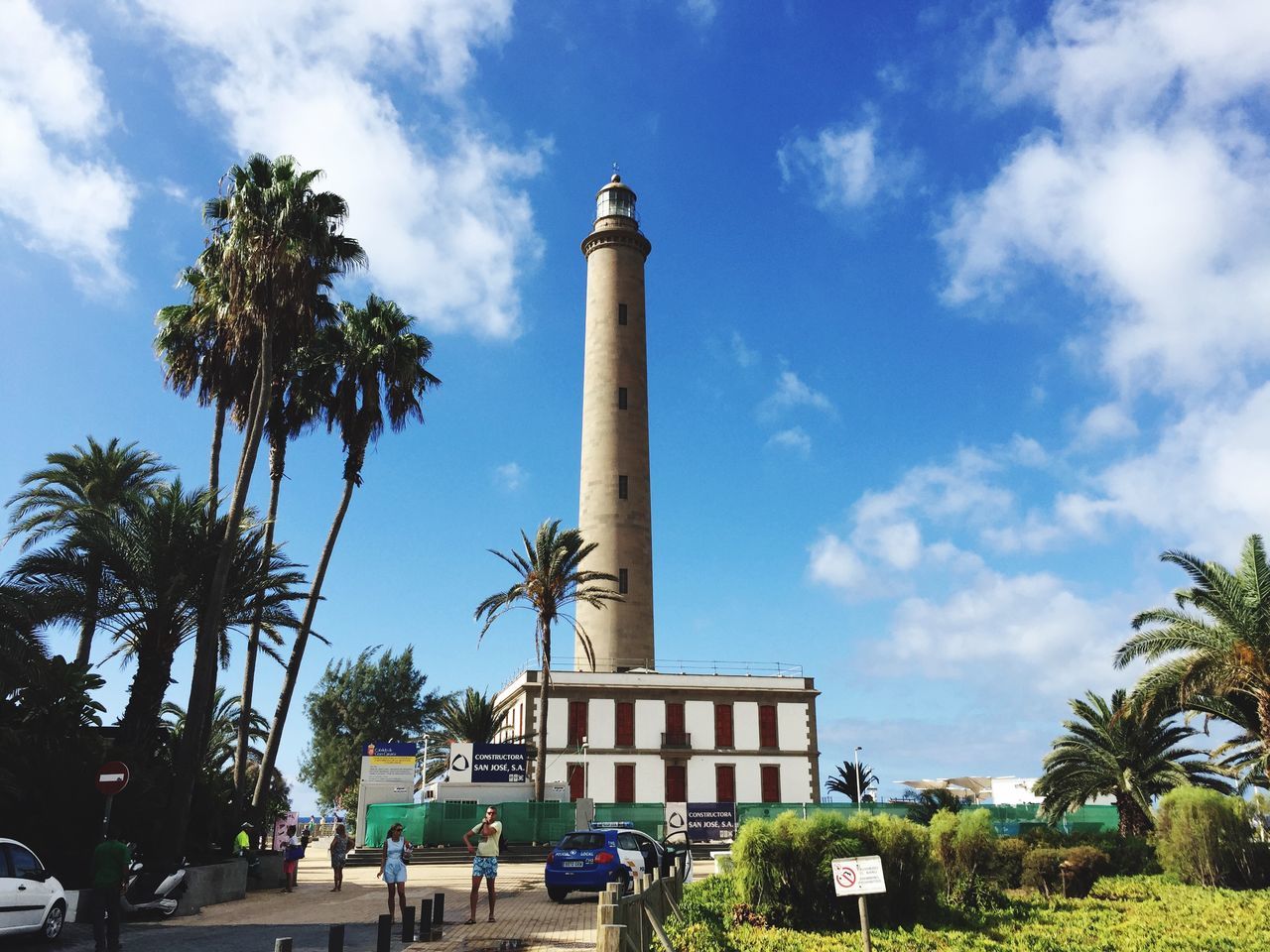 building exterior, architecture, built structure, sky, tree, guidance, low angle view, lighthouse, tower, cloud - sky, cloud, street light, palm tree, blue, direction, day, tall - high, safety, outdoors, incidental people