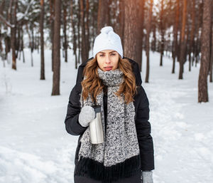 Portrait of confident woman holding insulated drink container while standing in forest during winter