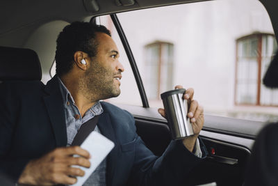 Smiling male commuter with insulated drink container and smart phone looking out through car window