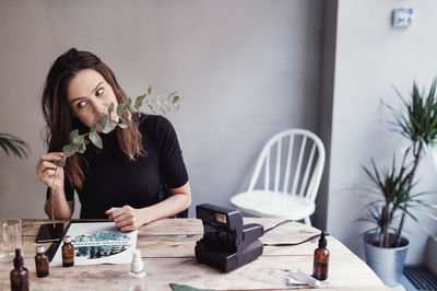 Woman smelling leaves while sitting at table against wall in perfume workshop