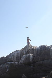 Low angle view of bird flying over shirtless man standing on rocks against clear sky