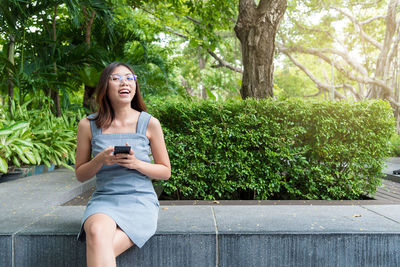 Smiling young woman using phone while sitting on tree