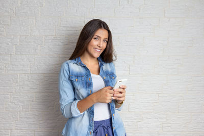 Portrait of smiling young woman holding smart phone while standing against wall