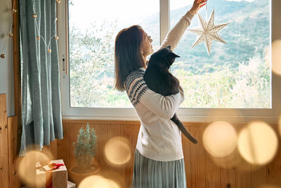 Woman in festive mood hugging black cat near the window while preparing for celebration christmas.