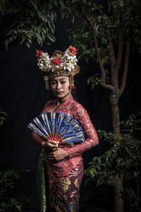 Portrait of woman in traditional clothing holding hand fan while standing by plants