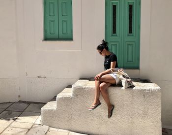 Young woman sitting on white staircase leading to the house with green door and shutters greece symi