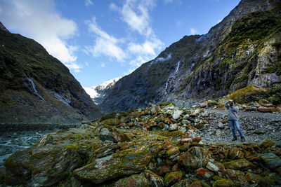 Man photographing while standing on rock by mountains against sky