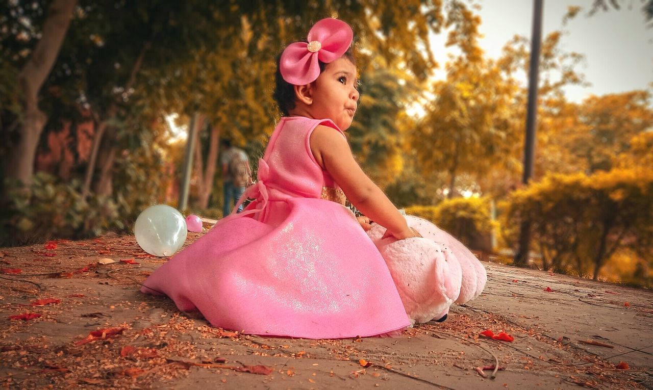 pink color, one person, real people, plant, full length, clothing, women, tree, leisure activity, nature, land, looking away, sitting, lifestyles, focus on foreground, childhood, side view, fashion, outdoors, innocence