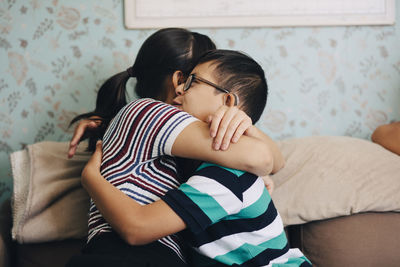 Sister embracing boy while sitting on sofa against wall at home