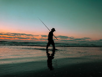 Silhouette man fishing at beach against sky during sunset